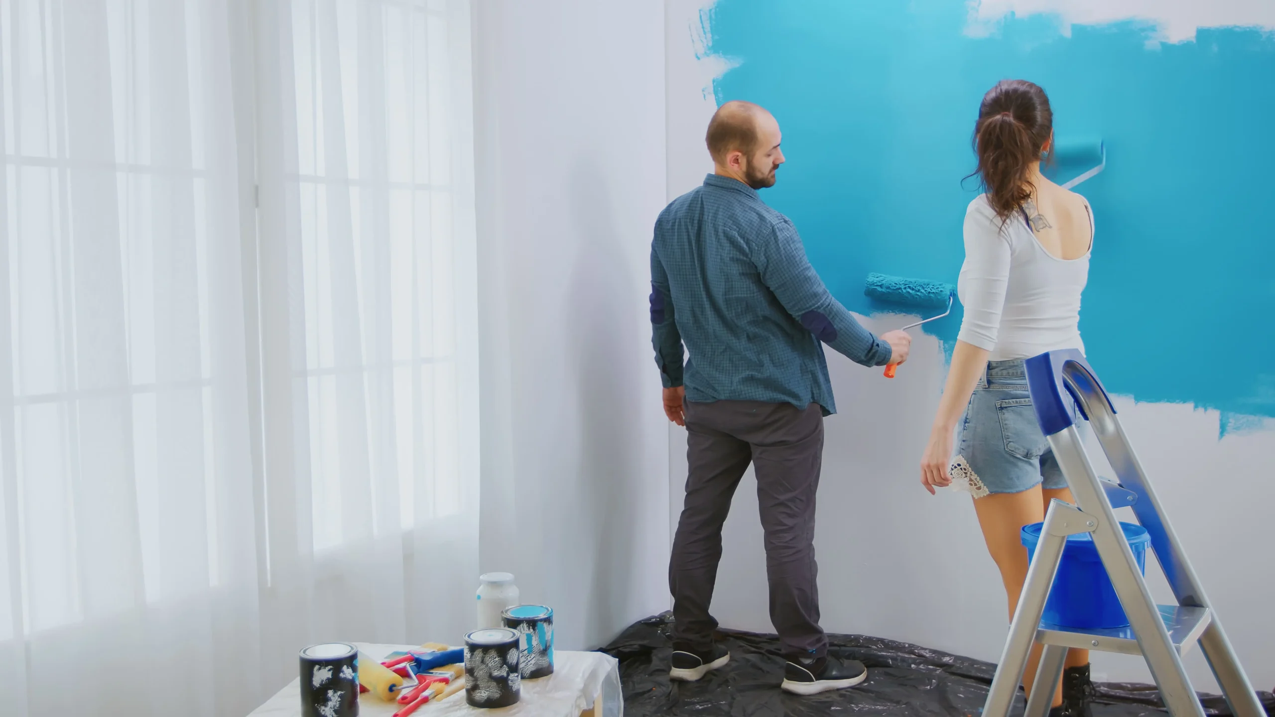caucasian couple painting wall with roller brush blue paint apartment redecoration home construction while renovating improving repair decorating 1 scaled